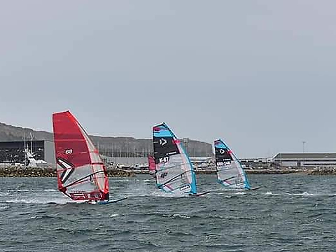 three windsurfers racing beneath pale blue sky, with backdrop of marina